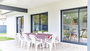 Ein Essbereich im Freien mit Tisch und Stühlen, großen Schiebeglastüren und Blick auf ein Haus im Hintergrund.