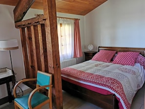 Un dormitorio con estructura de cama de madera, una silla con cojín verde, una mesita de noche con lámpara y una ventana con cortinas.