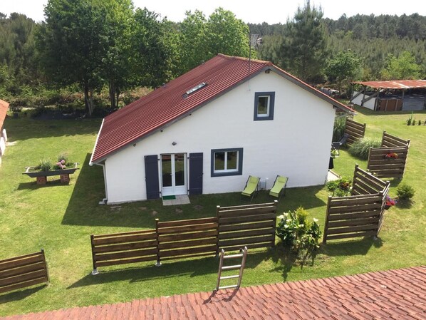 Ein weißes Gebäude mit rotem Dach, einem kleinen Fenster und einer Tür. Im Hof befinden sich Holzbänke und eine Leiter.