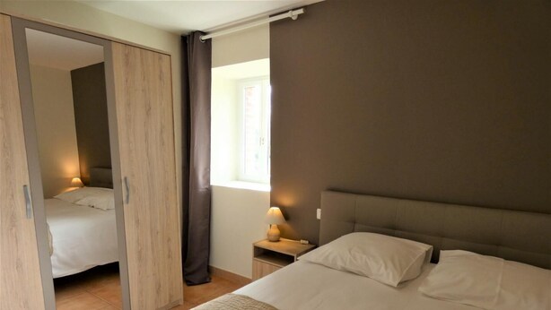 A hotel room with a bed, a nightstand with a lamp, a window with curtains, and a wardrobe with a mirror. - A Noust - Cagnotte - Gîte (CAGNOTTE)