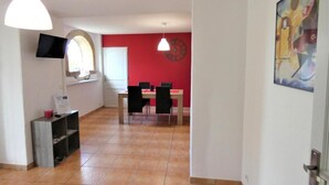 A spacious room with a tiled floor, a dining area with a table and chairs, a TV mounted on the wall, and a door with a handle. - A Noust - Cagnotte - Gîte (CAGNOTTE)