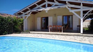 A covered outdoor area with a pool, wooden furniture, and a building with windows and doors.