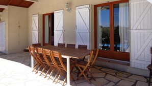 A covered outdoor dining area with a wooden table and chairs, white shutters, and a stone floor.