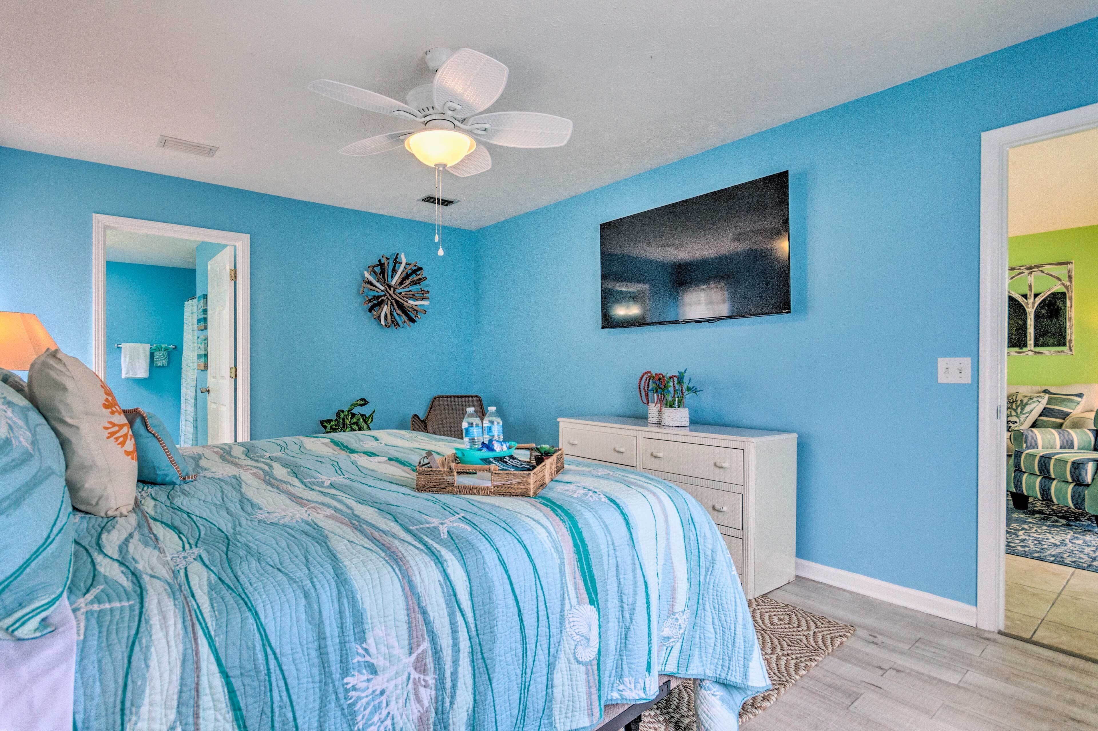 A bedroom with a bed, a dresser, a ceiling fan, a wall-mounted TV, and a door leading to another room.