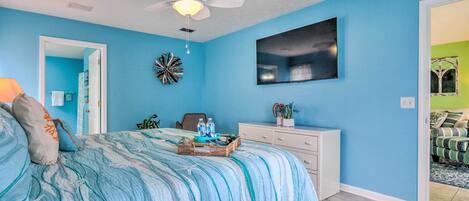 A bedroom with a bed, a dresser, a ceiling fan, a wall-mounted TV, and a door leading to another room.