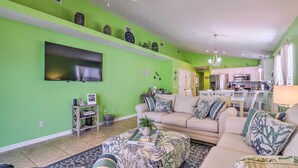 A living room with a green wall, a flat-screen TV, a sofa, a coffee table, and a kitchen in the background. - PCB Retreat w/ Patio & Grill < 1 Mi to Beach! (Panama City Beach)
