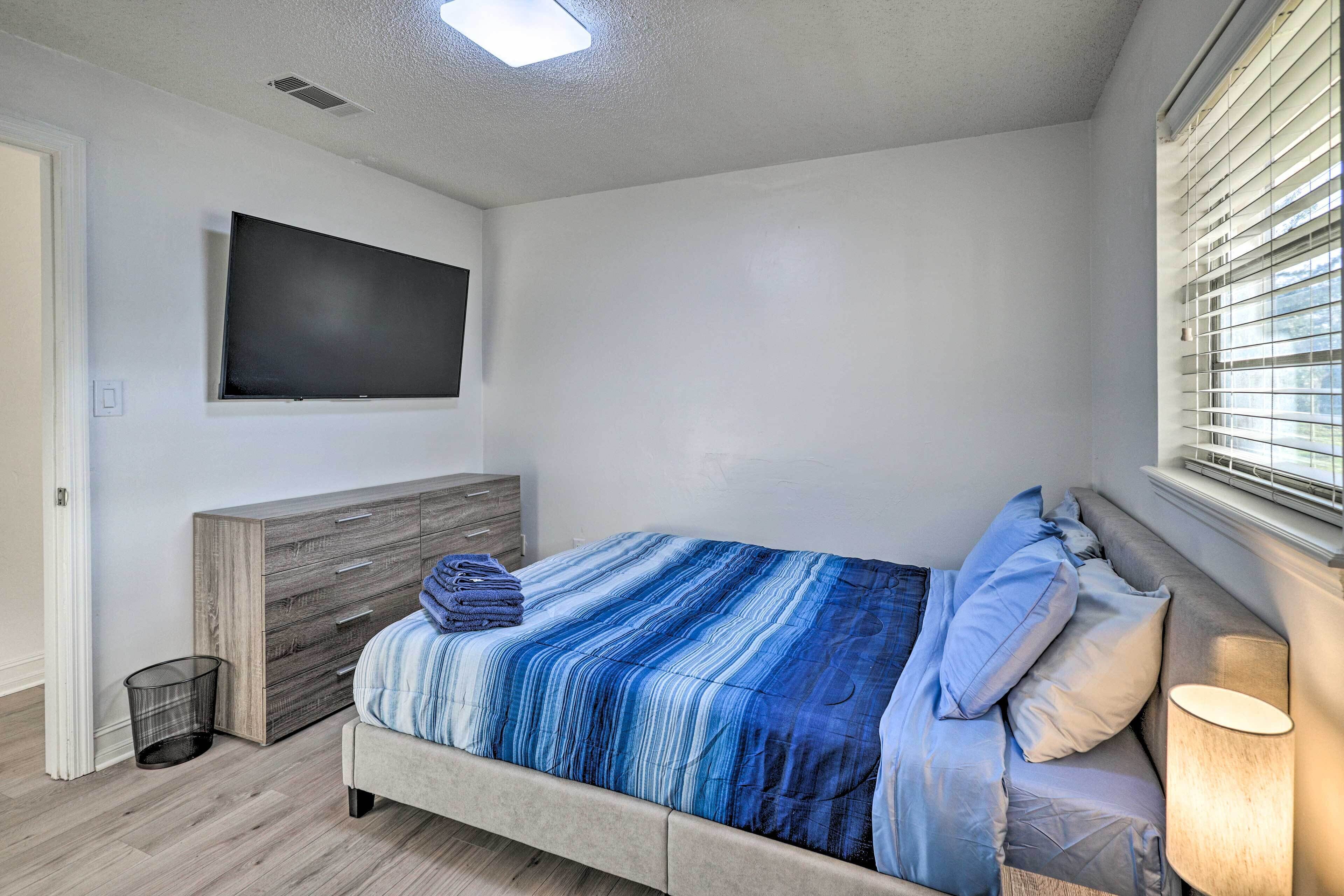 A bedroom with a bed, a TV mounted on the wall, a bedside table with a lamp, and a window with blinds.