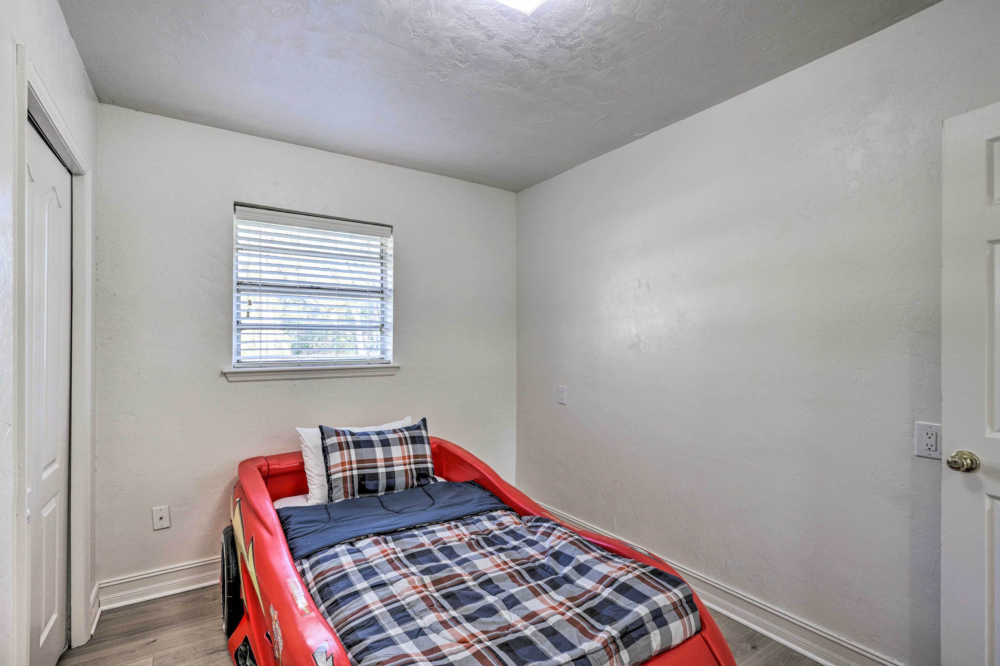 A small, single-bed room with a red bed frame, a window with blinds, and white walls.