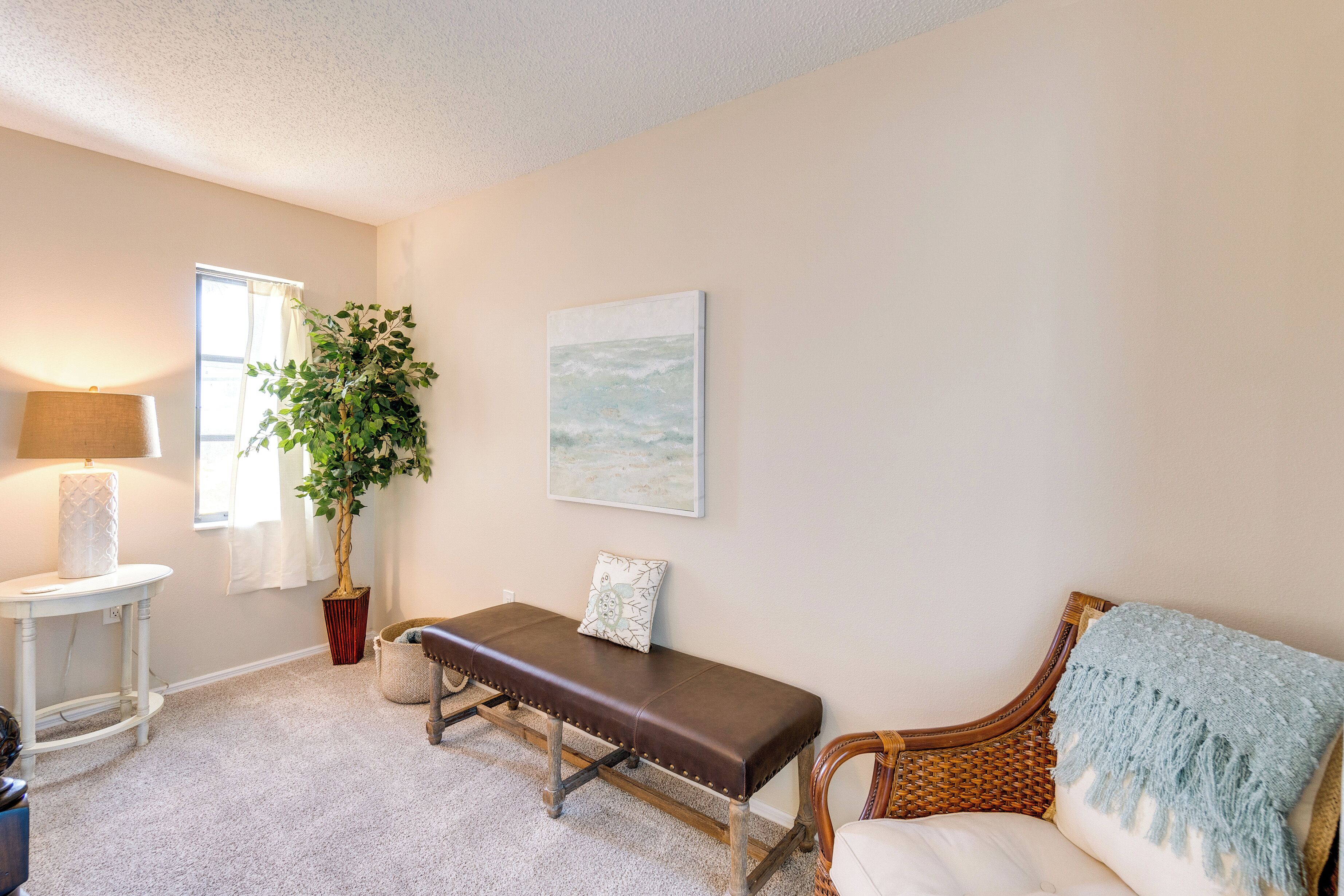 A room with a bench, a wicker chair, a lamp, a plant, and a painting on the wall.