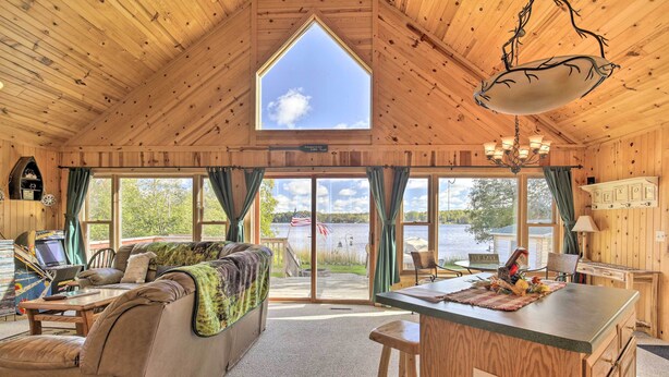 A cozy cabin with a large window, a dining area, and a sofa with a view of the lake. - Hurley Lakefront Hideaway w/ Fire Pit & Dock! (Hurley)