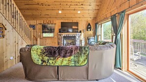 A cozy cabin interior with a wooden ceiling, a fireplace, a sofa with a blanket, and large windows. - Hurley Lakefront Hideaway w/ Fire Pit & Dock! (Hurley)