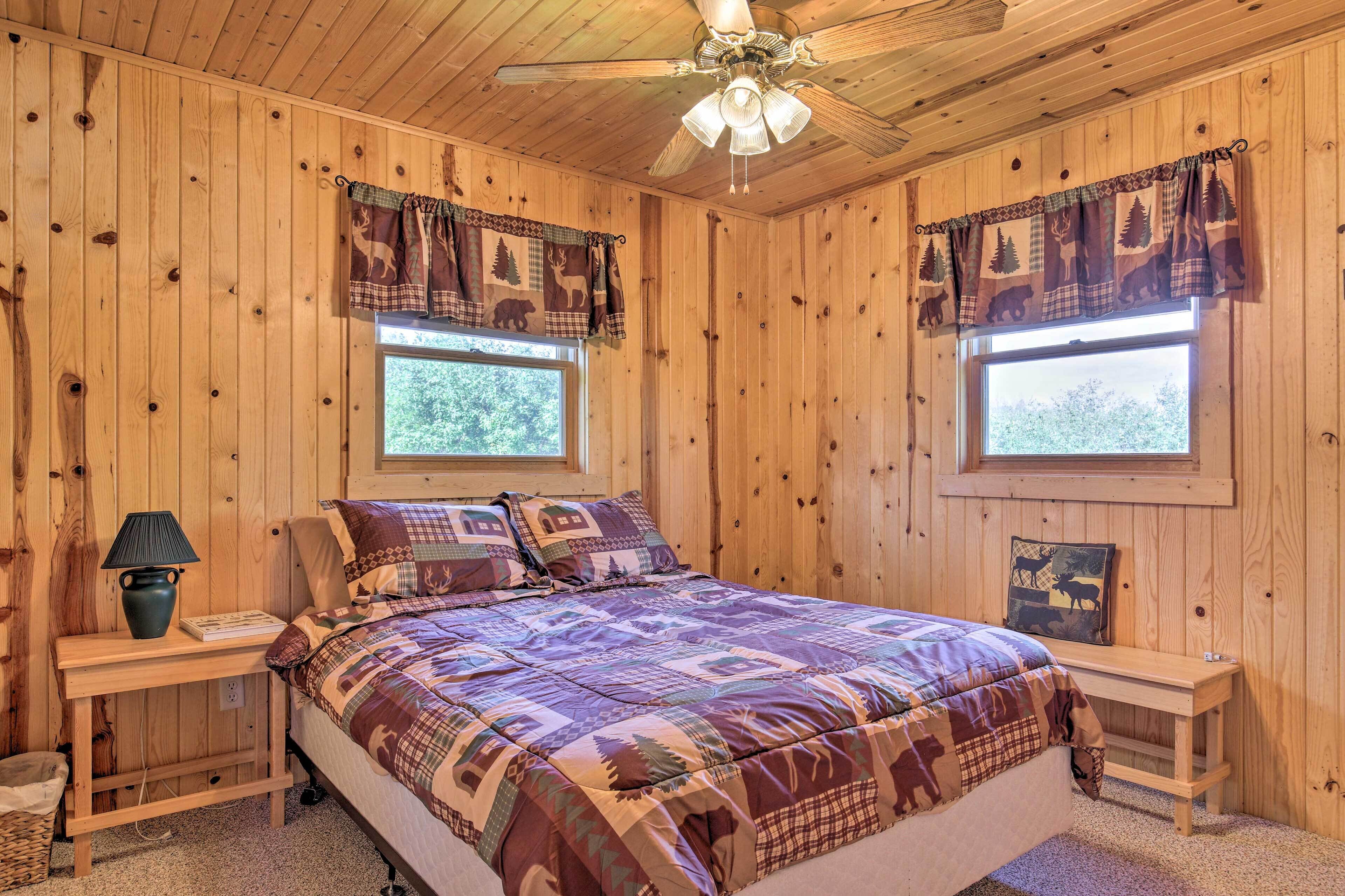 Une chambre à coucher dans une cabane en bois, avec un lit, deux fenêtres, un ventilateur de plafond et une table de chevet avec une lampe.