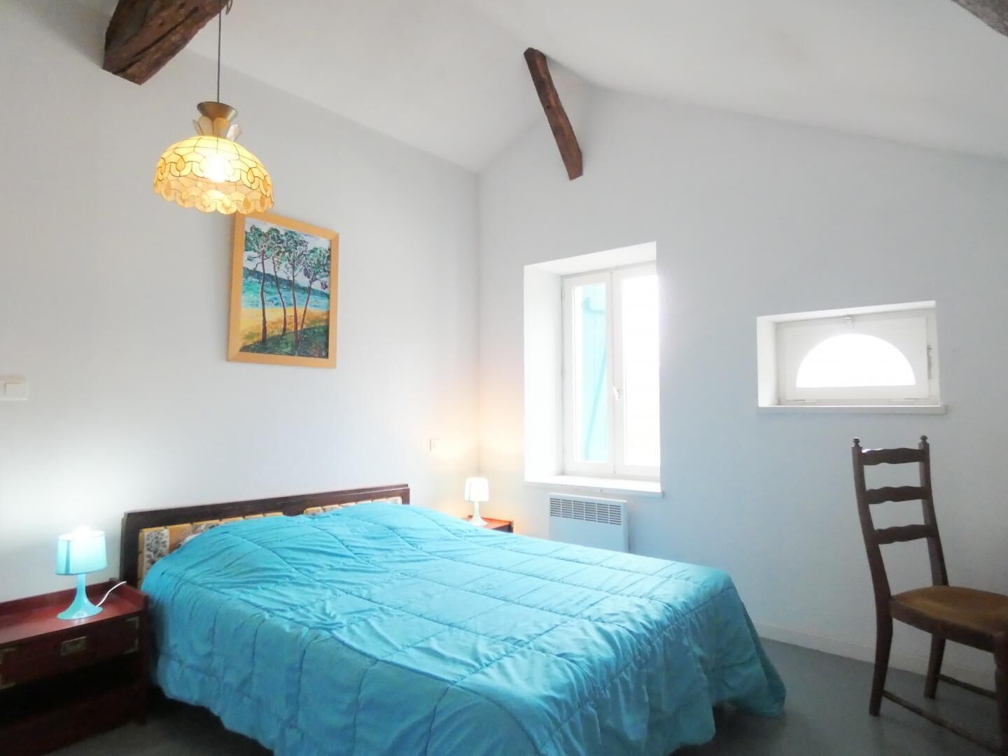 A bedroom with a bed, a chair, a window, a painting, and a hanging light fixture.