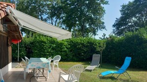 En udendørs terrasse med et bord og stole, en blå liggestol og et overdækket område med en rød parasol.