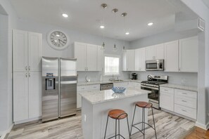 A modern kitchen with white cabinetry, stainless steel appliances, a central island with stools, and a clock on the wall. - 4 Blocks to Beach! Family-friendly Galveston Home (Galveston)
