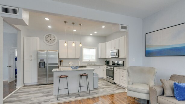 A modern kitchen with white cabinets, a stainless steel refrigerator, a microwave, and a stove. There is a large island with stools, and a beige armchair and couch in the living area. - 4 Blocks to Beach! Family-friendly Galveston Home (Galveston)