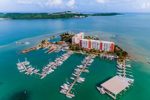 Une vue aérienne d’une marina où sont amarrés de nombreux bateaux, d’un grand bâtiment d’hôtel et qui offre une vue dégagée sur la côte environnante.