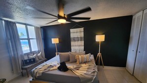 Et moderne hotelværelse med en stor seng, to sengeborde med lamper, ventilator i loftet og udsigt til havet, samt en væg med sort detalje.