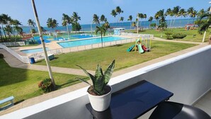 En balkon med en potteplante, to sorte stole og udsigt til en pool og strand.