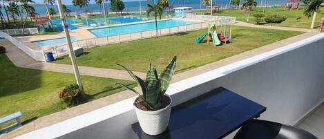 En balkong med en krukväxt, två svarta stolar och utsikt över en pool och en strand.