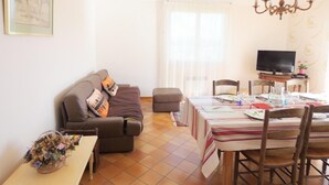 A living room with a sofa, a dining table set for two, a television, and a chandelier. - La Tanière - St-martin-de-seignanx (ST-MARTIN-DE-SEIGNANX)
