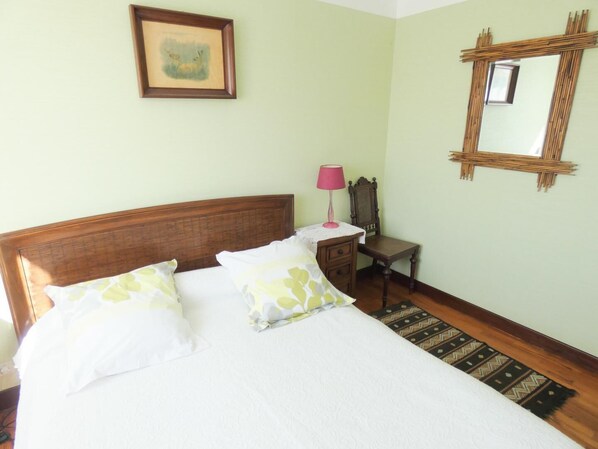 A neatly made bed with white linens and decorative pillows, a bedside table with a lamp, a chair, a framed picture on the wall, and a mirror with a bamboo frame.