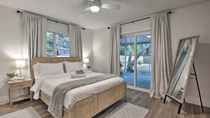 A bedroom with a bed, bedside tables, a ceiling fan, a large window with curtains, and a mirror on a stand.