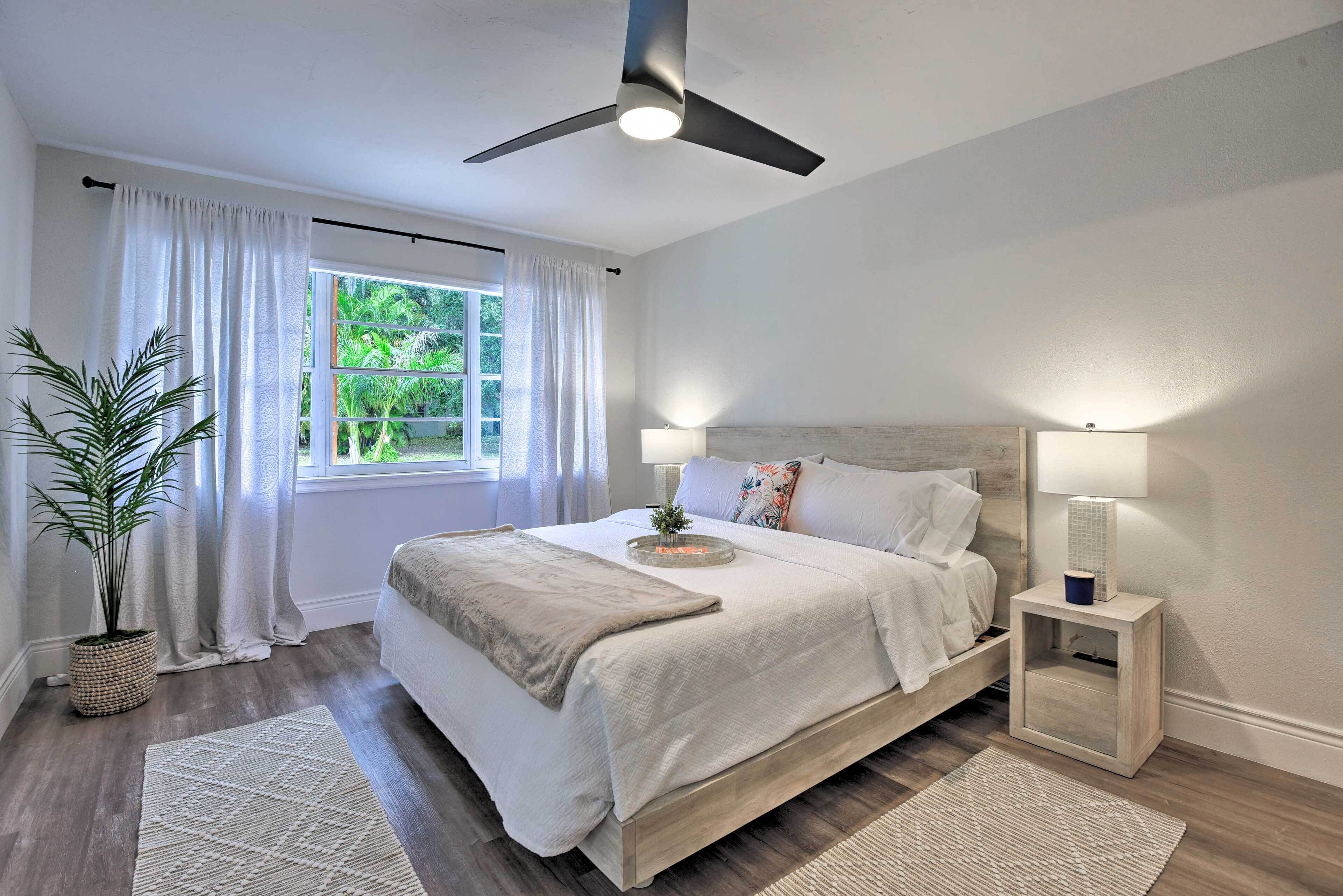 A bedroom with a bed, bedside lamps, a ceiling fan, a window with curtains, and a potted plant.