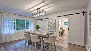 A dining area with a rectangular table, four chairs, a chandelier, and a view of a garden through large windows.
