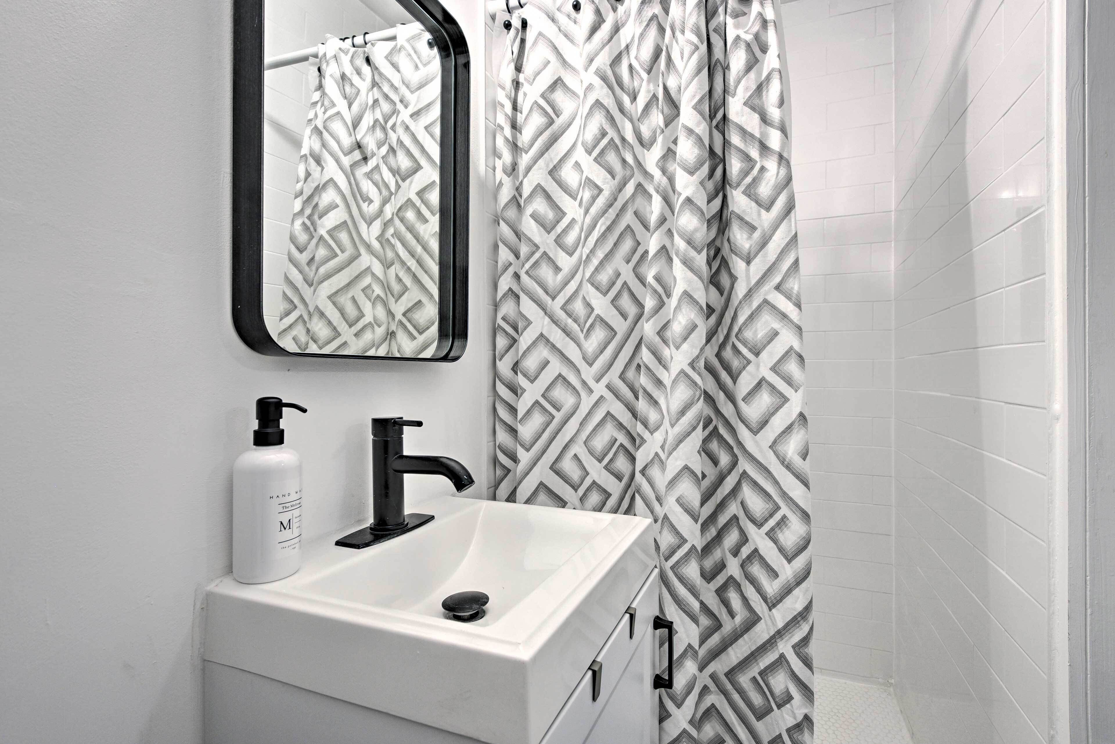 A bathroom with a white sink, black faucet, and a mirror above it. There is a patterned shower curtain and white tiled walls.