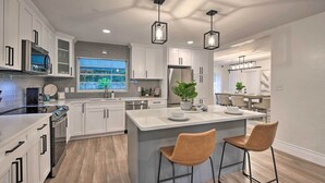 A modern kitchen with white cabinetry, a central island with two bar stools, a stainless steel refrigerator, and pendant lighting.