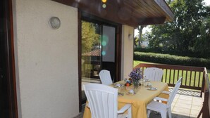 An outdoor patio with a table set for four, surrounded by white chairs, a yellow tablecloth, and a vase of flowers.