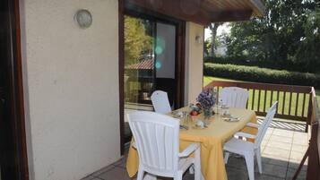 An outdoor patio with a table set for four, surrounded by white chairs, a yellow tablecloth, and a vase of flowers.