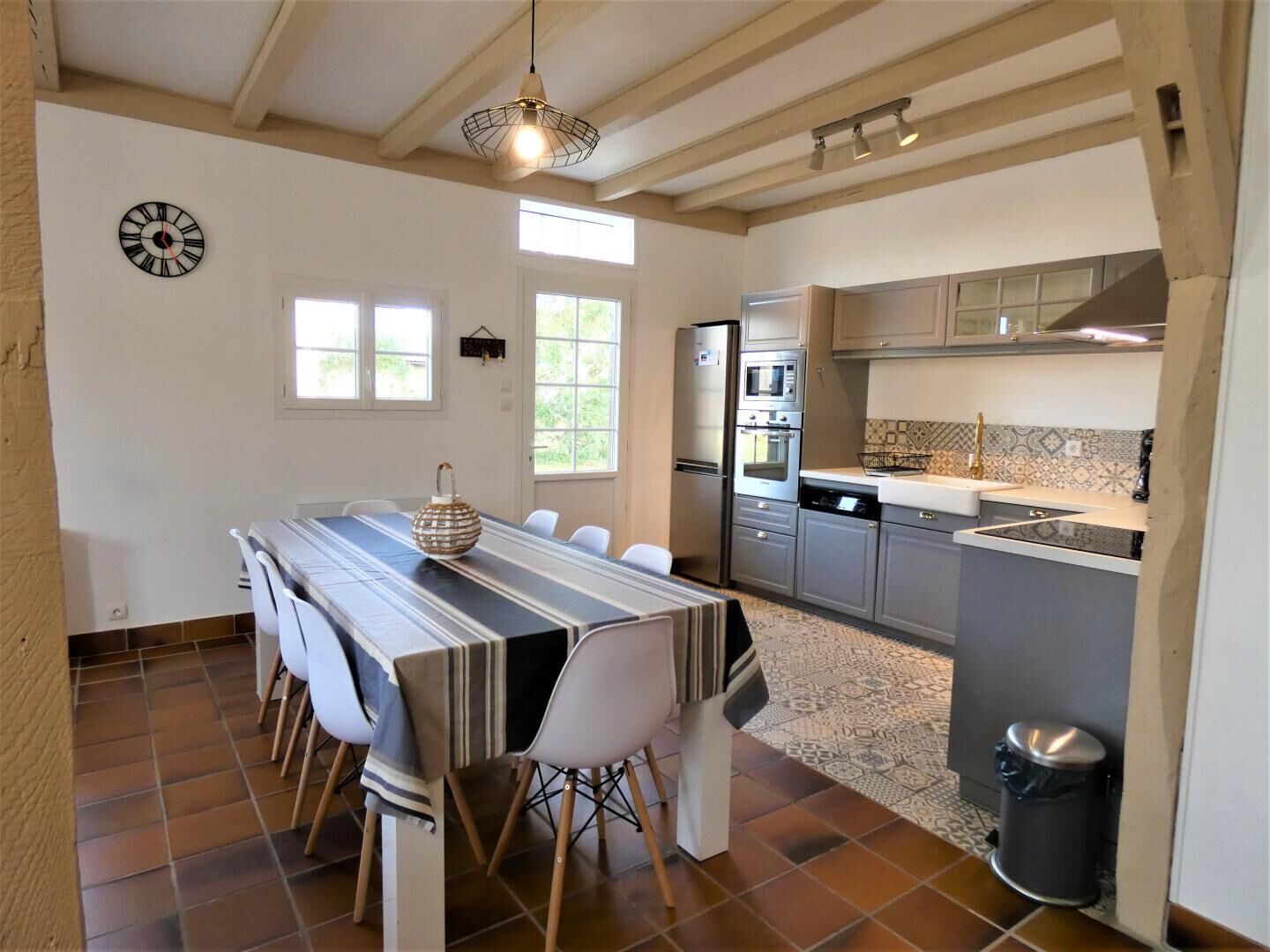 Un comedor con una mesa larga, sillas, un reloj y una cocina con electrodomésticos.