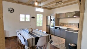 A dining area with a long table, chairs, a clock, and a kitchen with appliances.