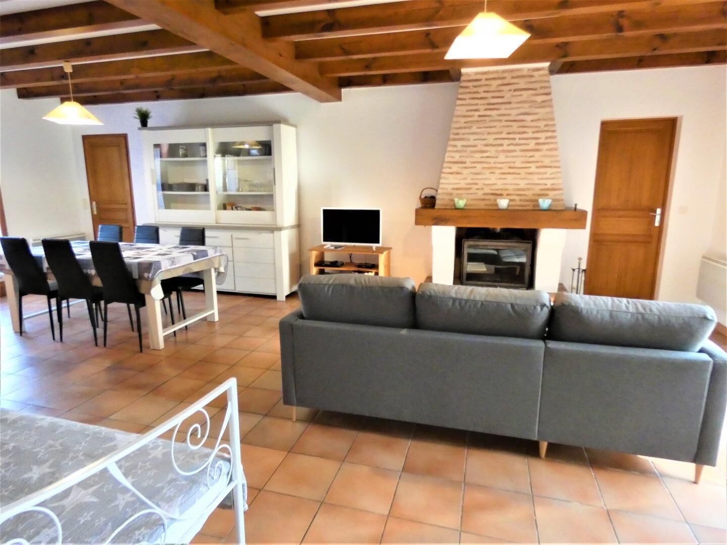 A living room with a grey sofa, wooden ceiling beams, a fireplace, and a dining area with a table and chairs.