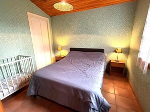 A bedroom with a bed, two bedside tables with lamps, a white door, and a crib. - La Prairie - Leon - Gîte (LEON)