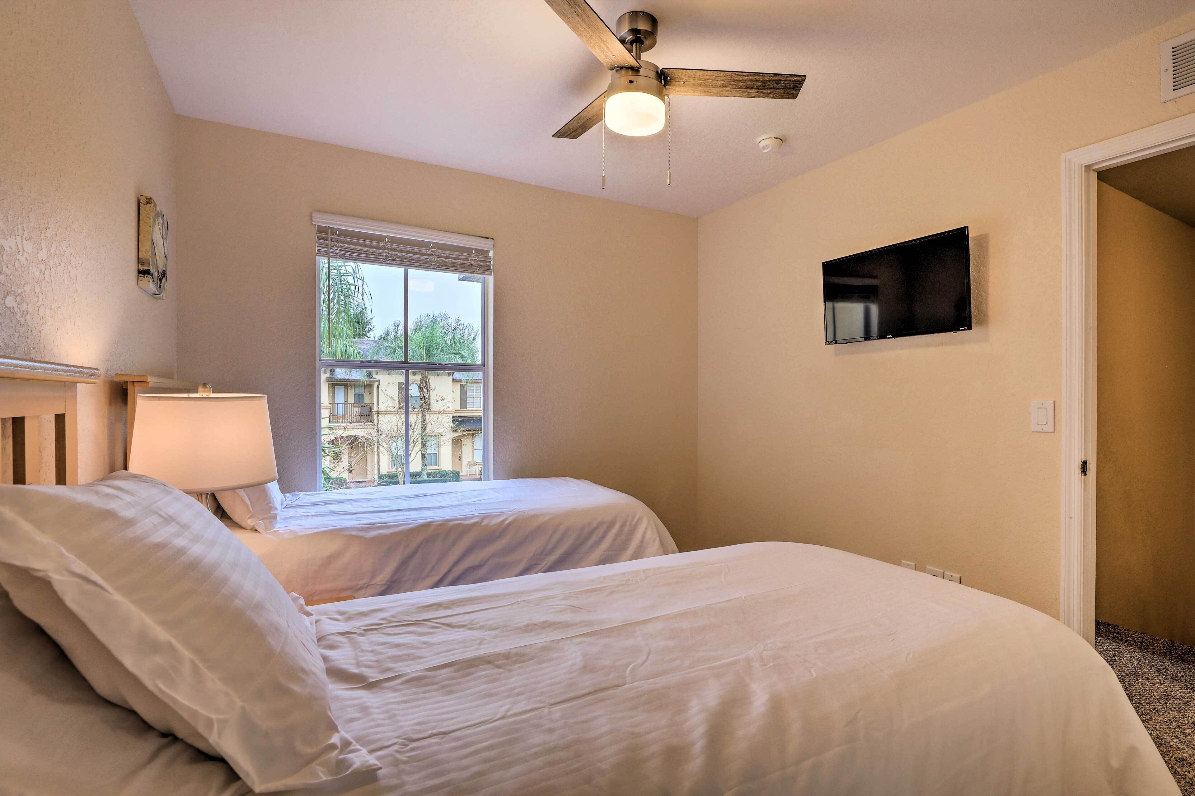 A bedroom with two beds, a ceiling fan, a window with blinds, a TV mounted on the wall, and a door leading to another room.