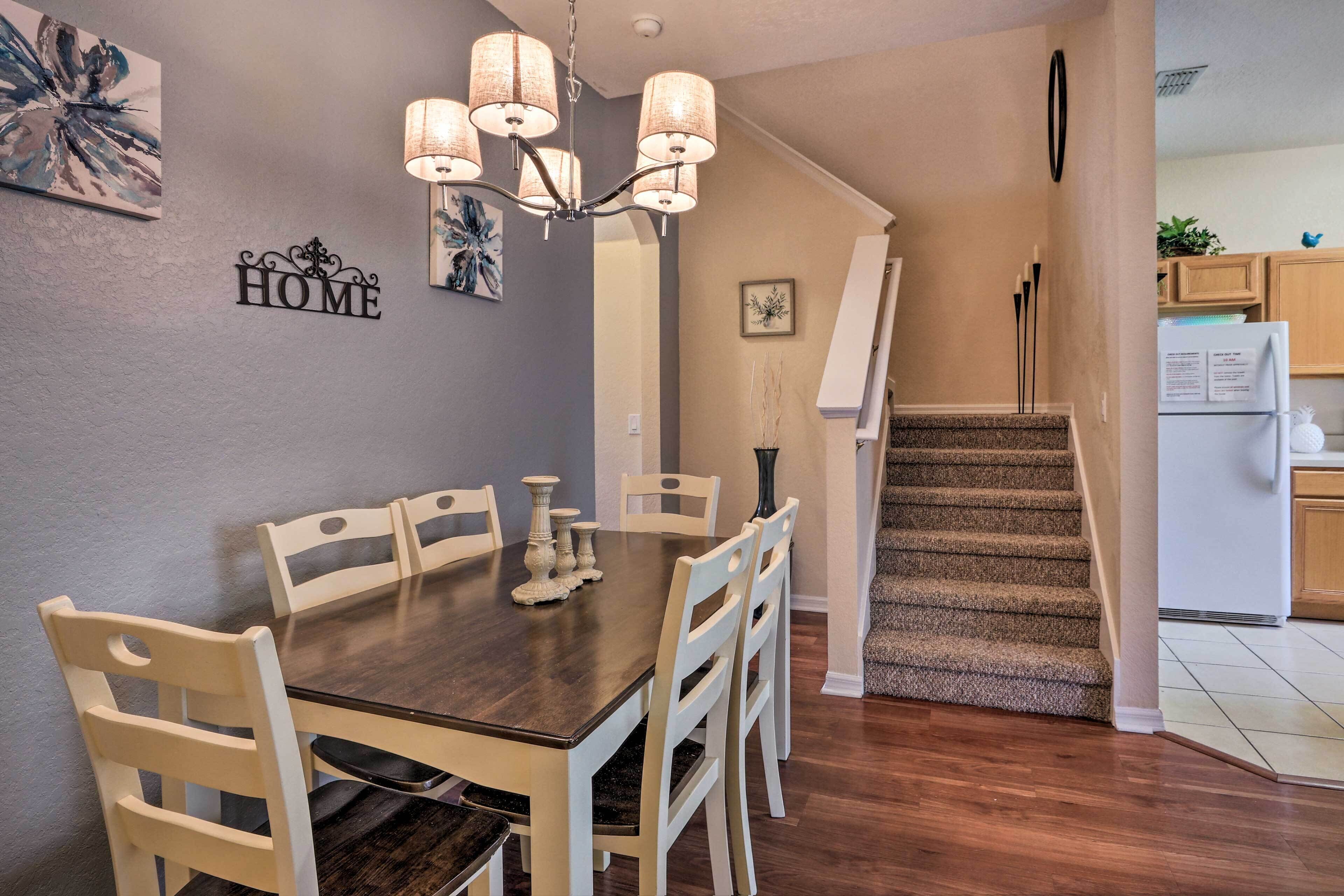 A dining area with a dark wooden table and white chairs, a chandelier, and a staircase leading to a kitchen.