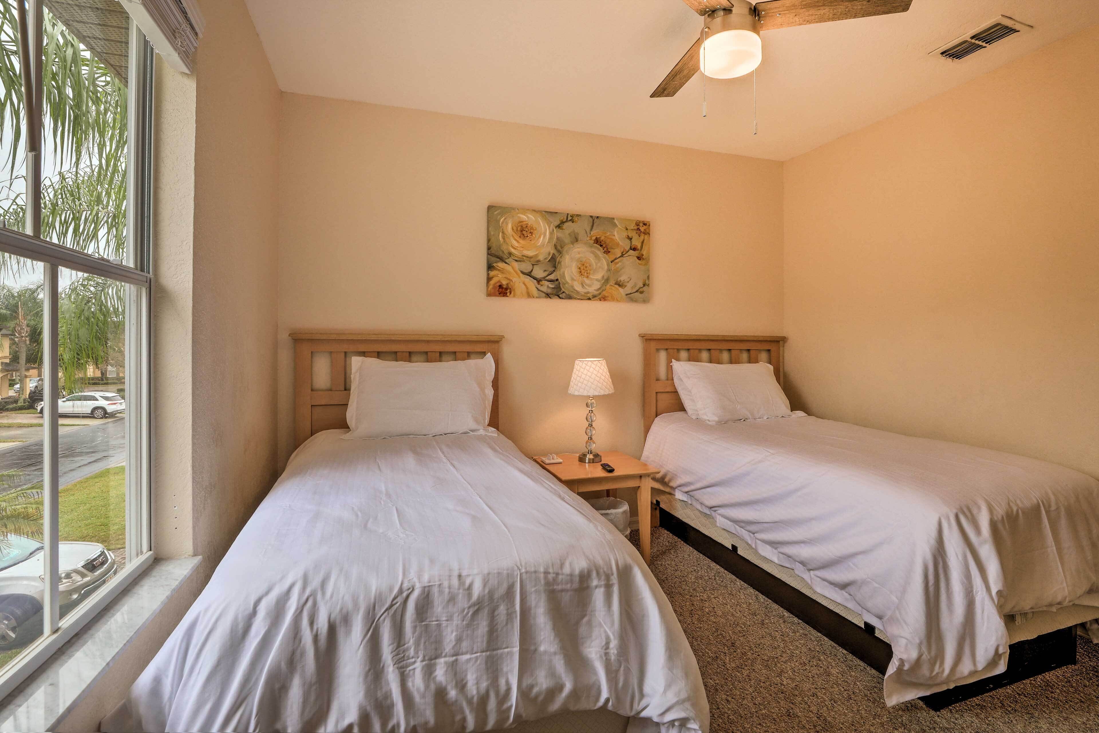 A hotel room with two beds, a ceiling fan, a window with a view of the street, and a floral painting on the wall.