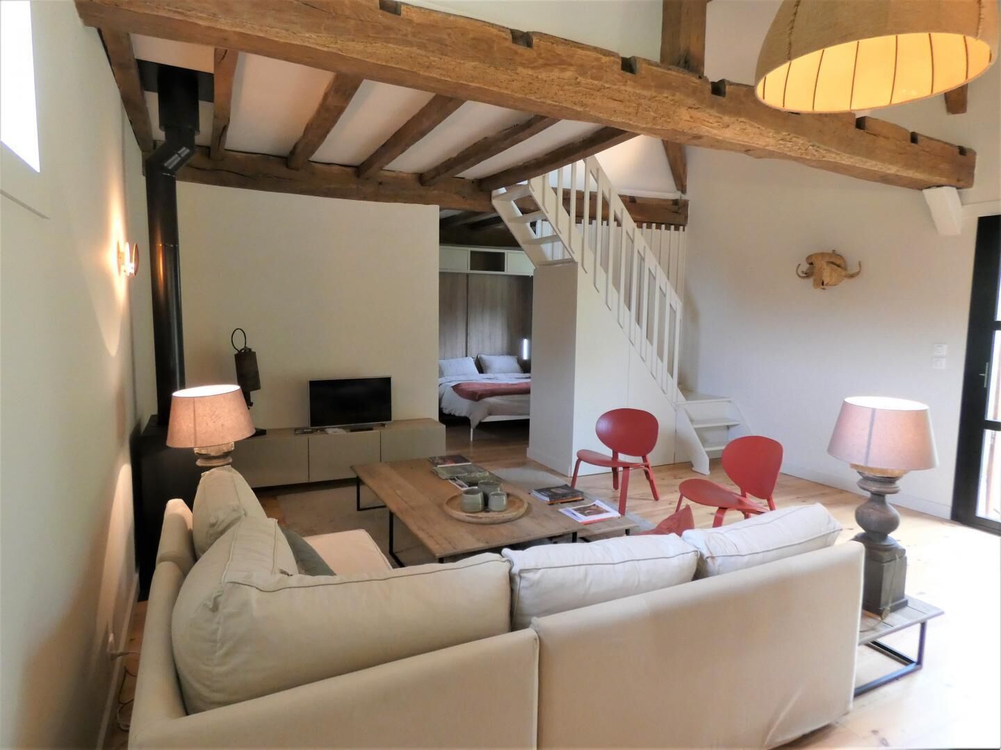A cozy living room with a wooden ceiling, a sofa, a coffee table, and a TV. A staircase leads to another room.