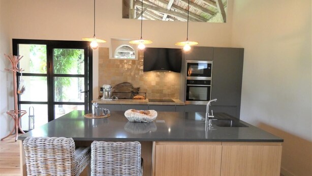 A modern kitchen with a large island, two wicker chairs, a built-in oven, and pendant lighting. - Les Ecuries du Château (BIAUDOS)
