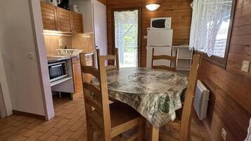 A wooden cabin interior with a dining table, chairs, a kitchen area with appliances, and a window with curtains.