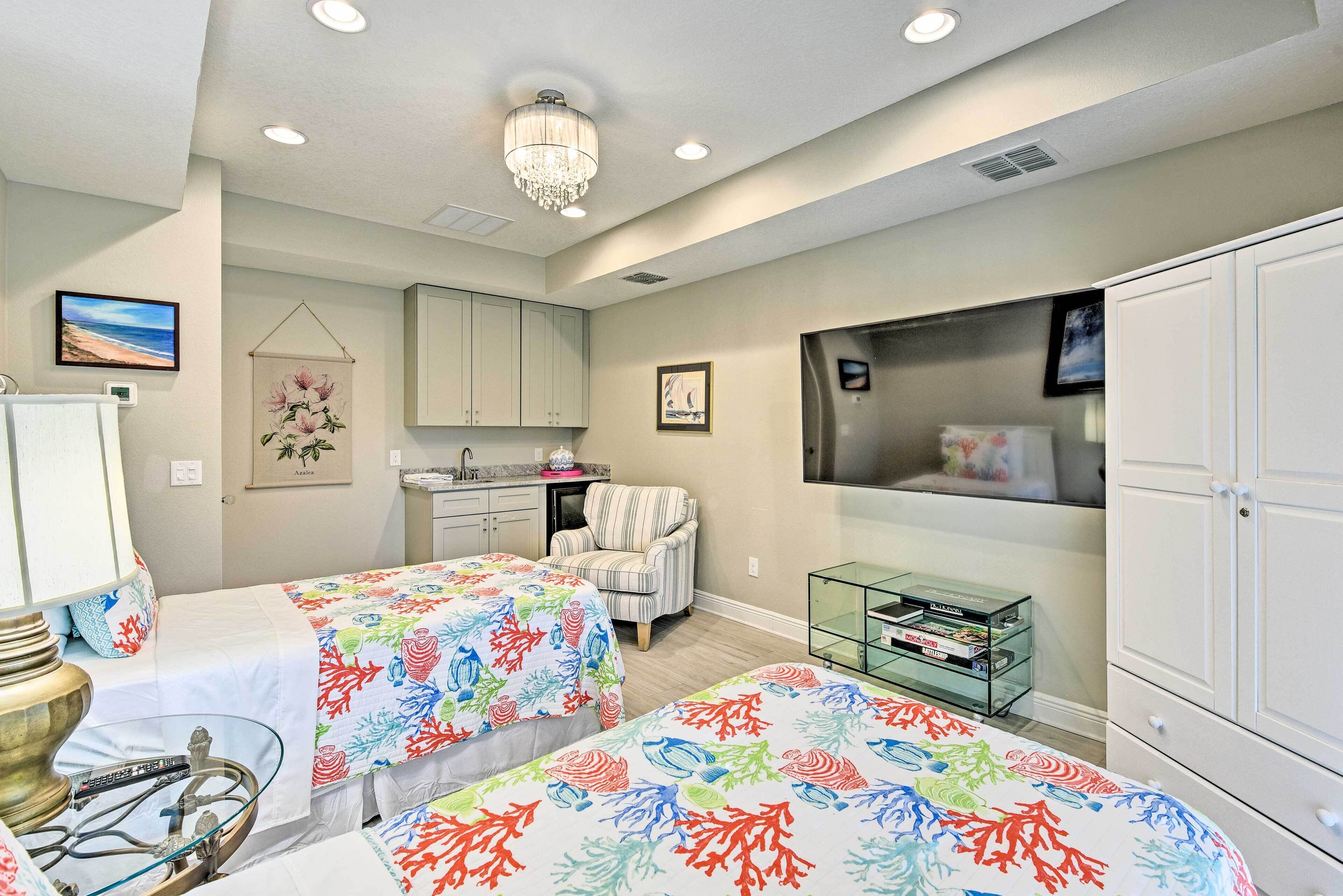 A bedroom with two beds, a chandelier, a TV, and a small kitchen area.