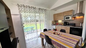 Une cuisine compacte dotée d’appareils électroménagers modernes, un coin repas avec une table dressée pour deux personnes, et une vue sur l’extérieur à travers un rideau à motifs.