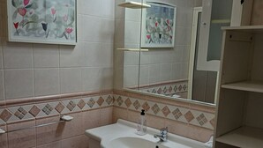 Une salle de bain avec un lavabo blanc, un miroir et des murs carrelés. Des carreaux décoratifs ornent le bas des murs et une étagère supporte une plante.
