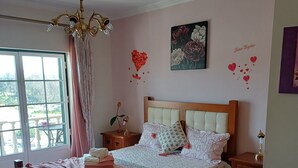 Une chambre à coucher avec un lit en bois, un lustre, une table de chevet, une commode, une fenêtre avec des rideaux et un mur orné d’un tableau floral et de décorations en forme de cœur.