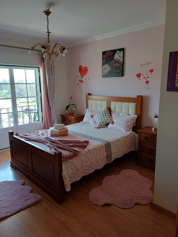 Un dormitorio con una cama de madera, una araña, una mesita de noche y cortinas en la ventana. En la pared, un cuadro floral y adornos con forma de corazón.