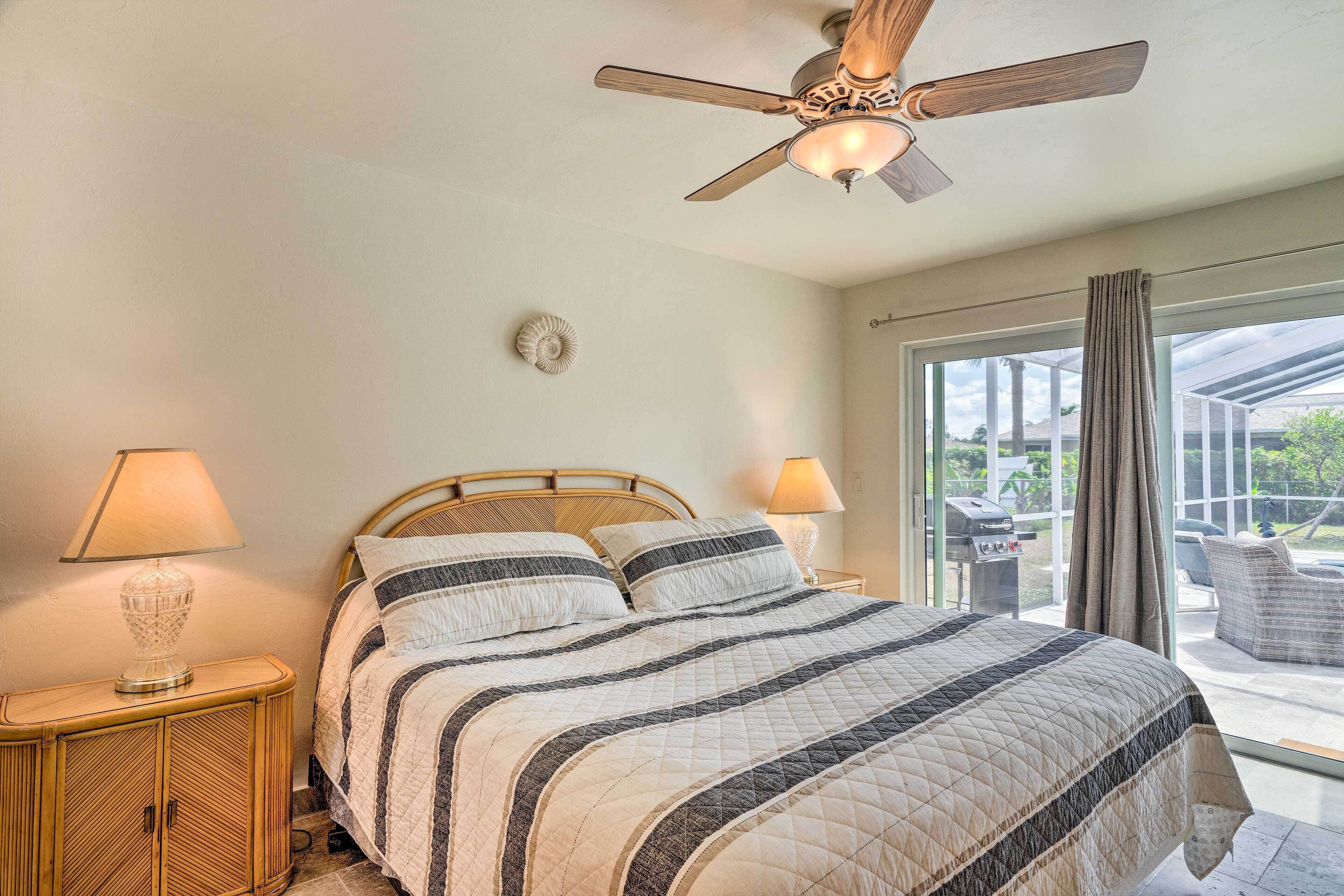 A bedroom with a bed, two bedside lamps, a ceiling fan, and a view of the outdoors through a large window.
