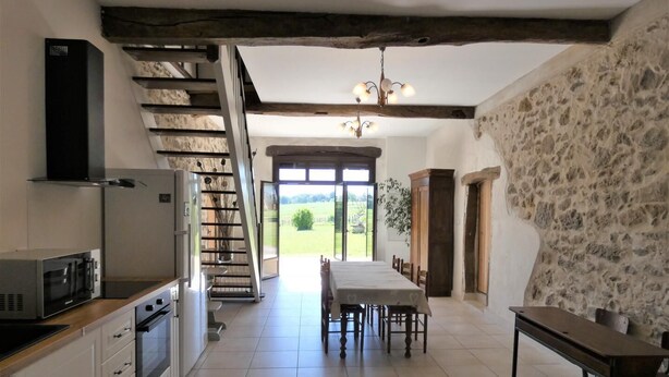 Eine Küche mit modernem Herd, einer Mikrowelle und einem eingebauten Backofen. Ein Esstisch mit Stühlen und eine Chandelieber befinden sich darüber. Eine Treppe führt zu einem Außenbereich mit Blick auf die Landschaft.